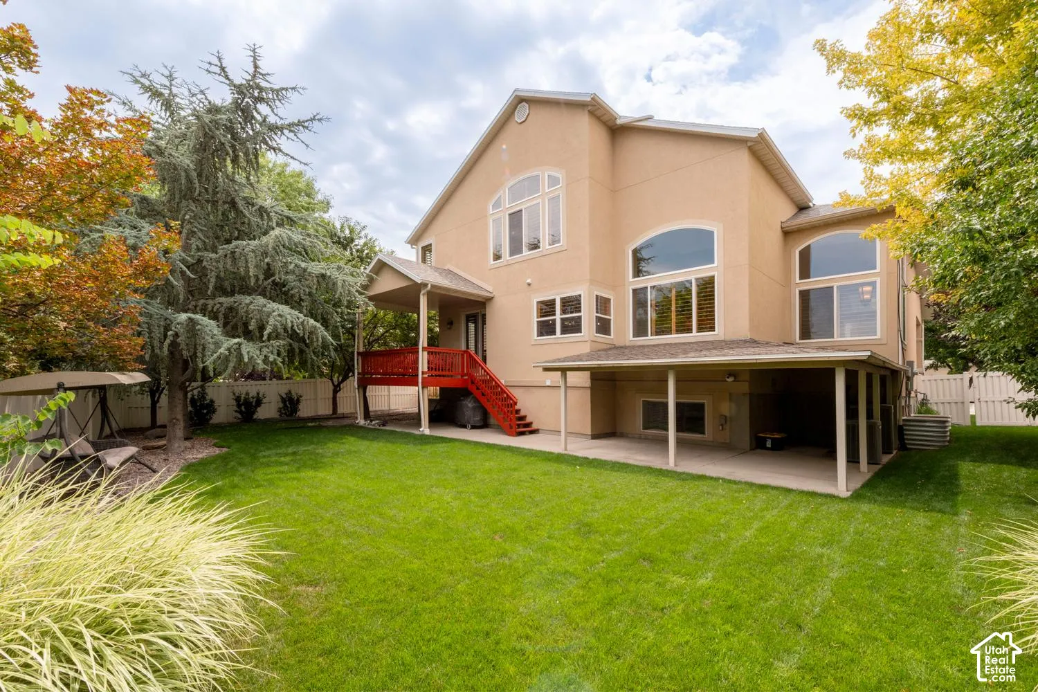 Expansive backyard featuring a manicured lawn, mature trees, a covered patio, and a covered deck with red-stained stairs
