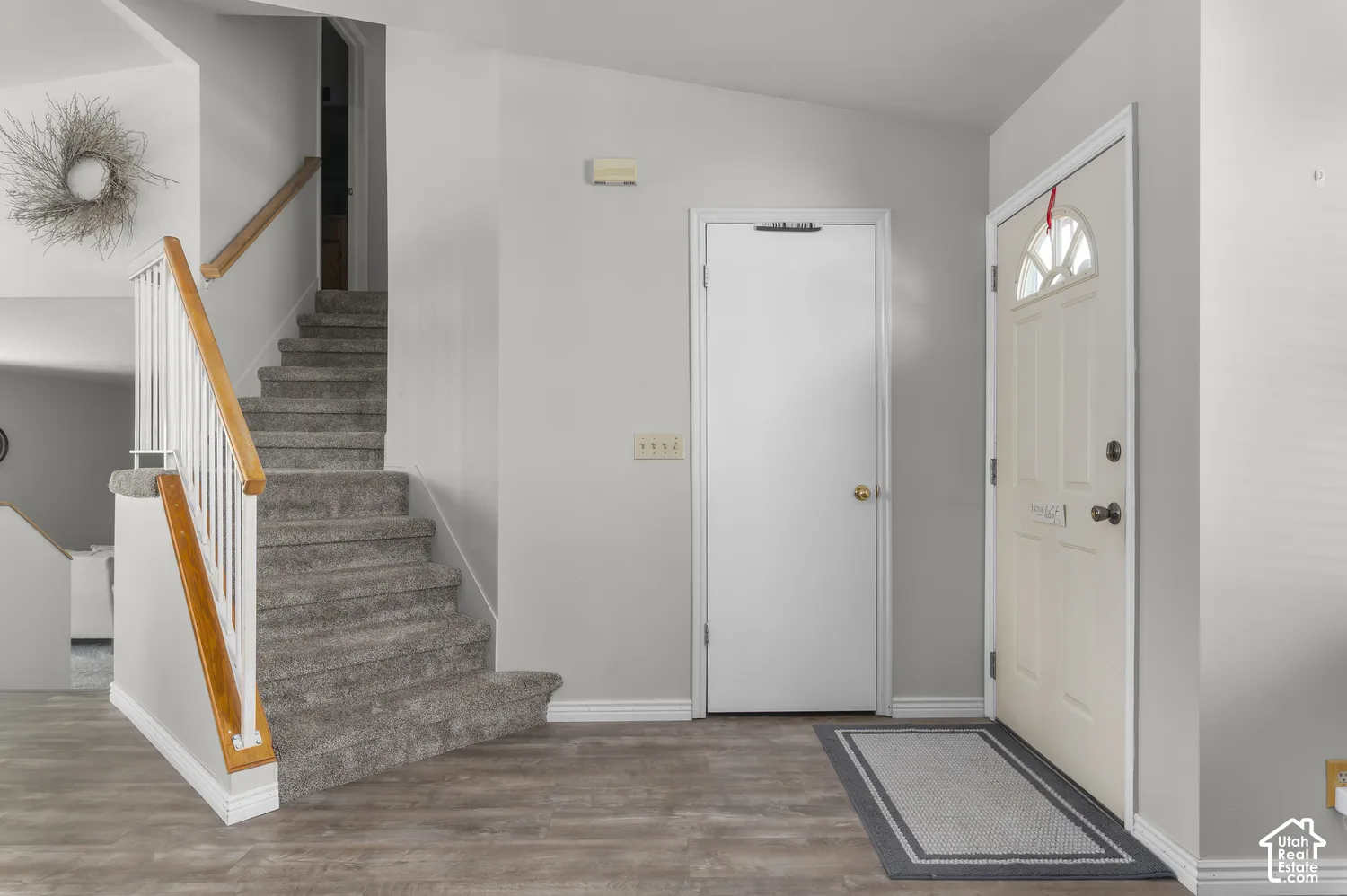 Wood-finish flooring, carpeted stairs with wood railing, white trim, and a front entry door with an arched glass insert