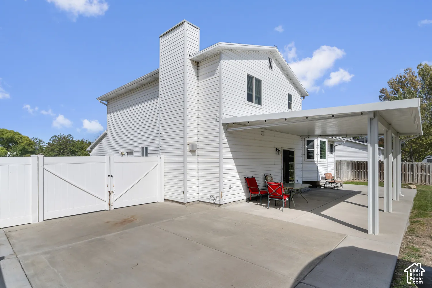 White siding exterior with a prominent chimney, covered patio, concrete patio area, and white privacy fencing with double gates