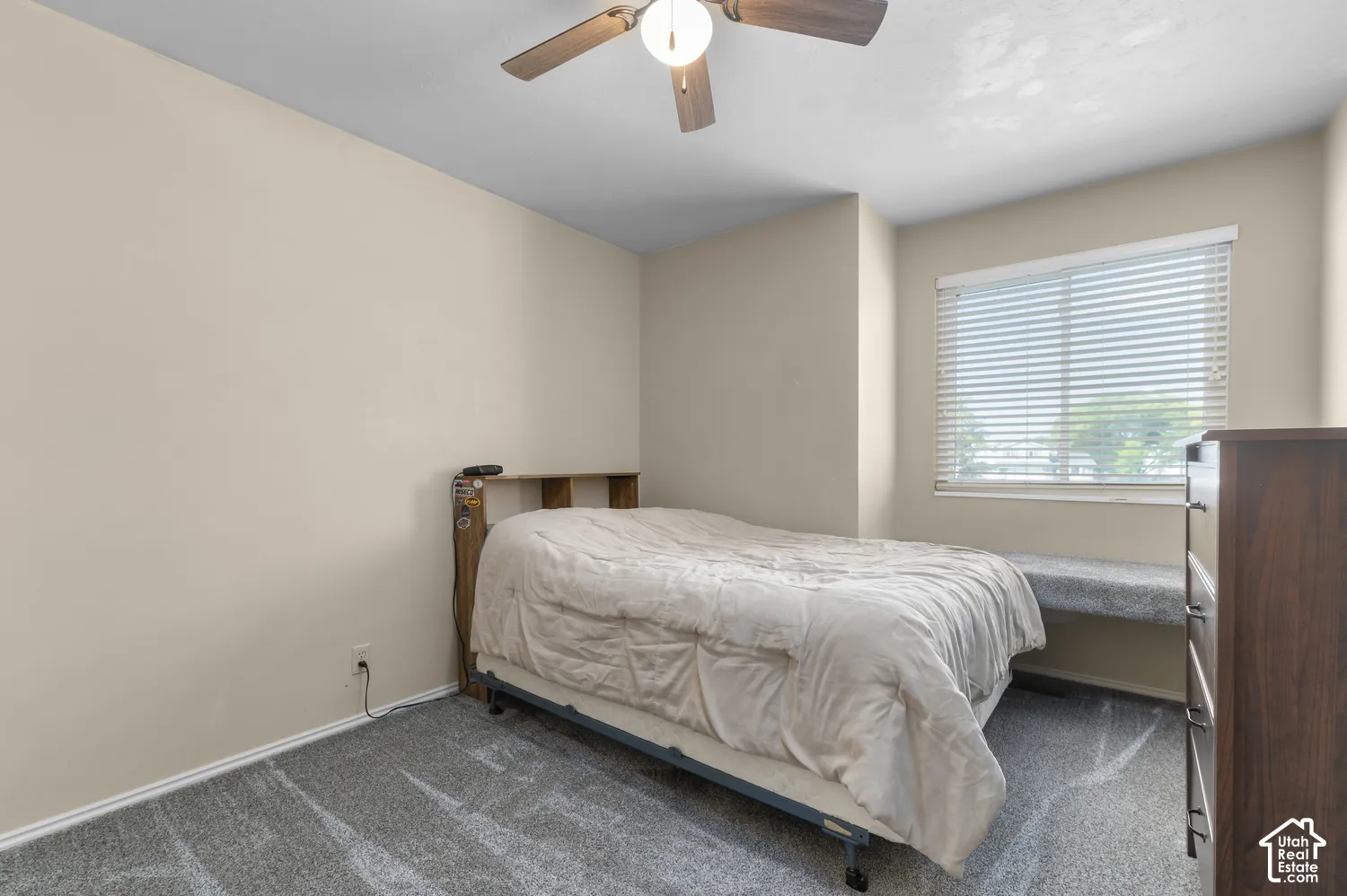 Plush carpet flooring, neutral wall paint, ceiling fan with integrated lighting, window with blinds, and a built-in window bench