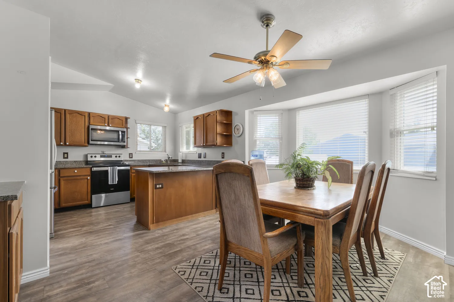 Kitchen and dining area featuring wood-finish flooring, wood cabinetry, stainless steel appliances, a center island with a sink, and a bay window