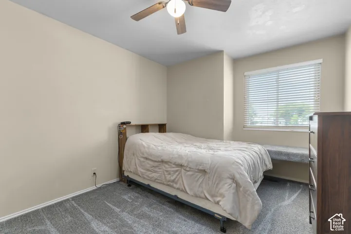 Plush carpet flooring, neutral wall paint, ceiling fan with integrated lighting, window with blinds, and a built-in window bench
