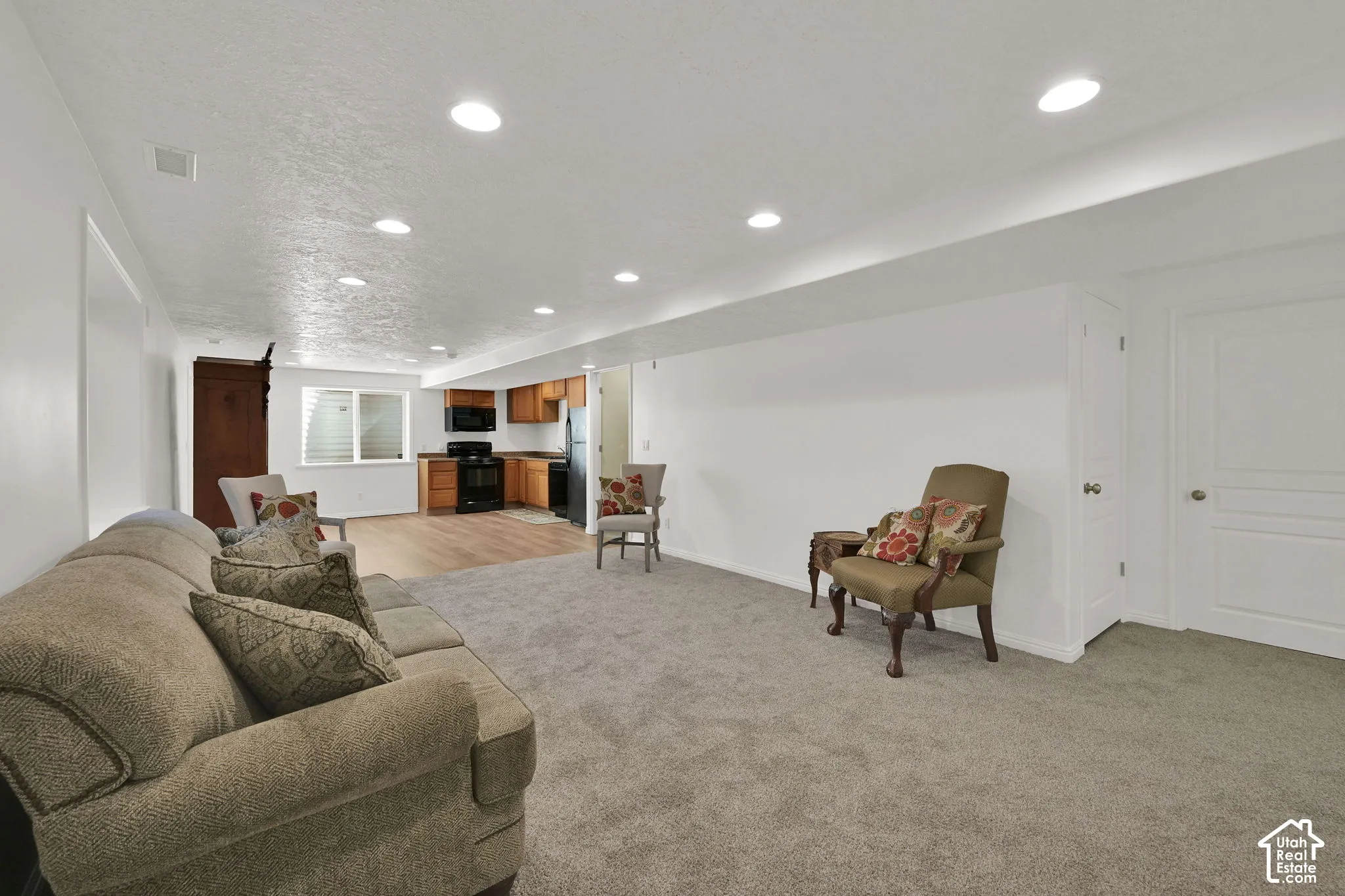 Recessed lighting illuminates the open-concept living space, featuring neutral carpeting and a kitchen with wood-finish cabinetry, a black range, and a black refrigerator