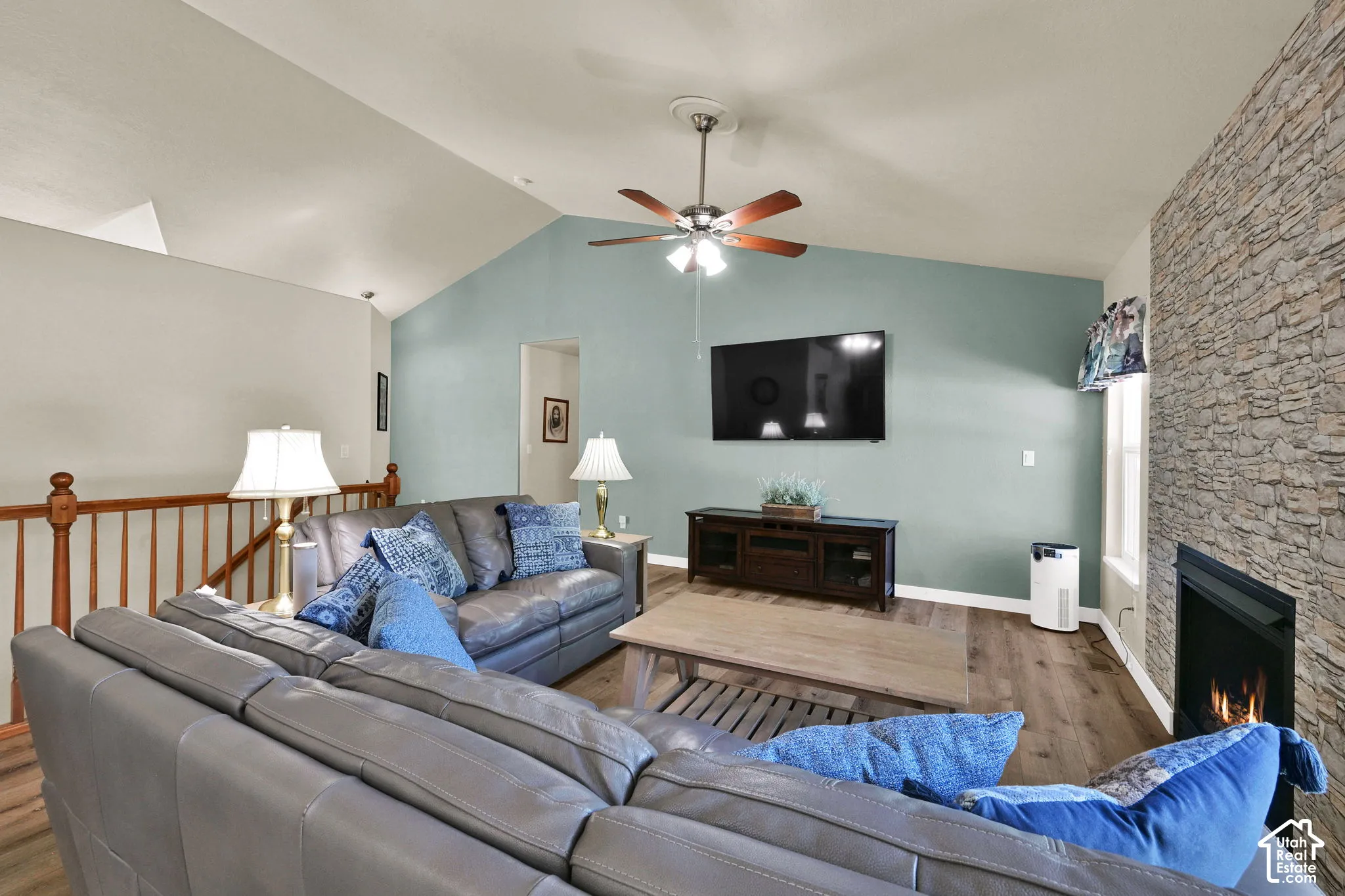 Vaulted ceiling, stone-clad fireplace, wood-finish flooring, ceiling fan, and wooden railing