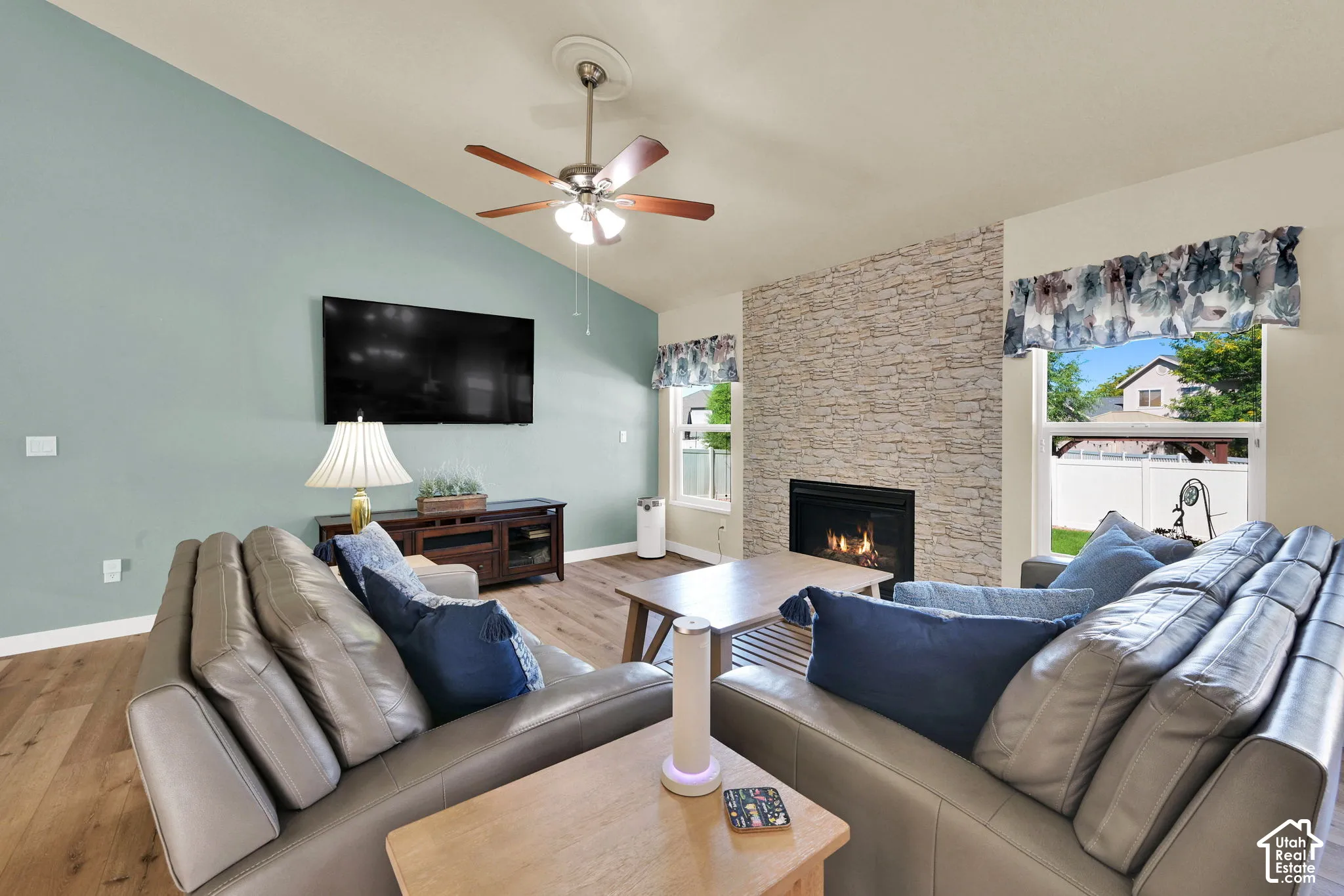 Vaulted ceiling with a ceiling fan, gas fireplace with a stone surround, and wood-finish flooring