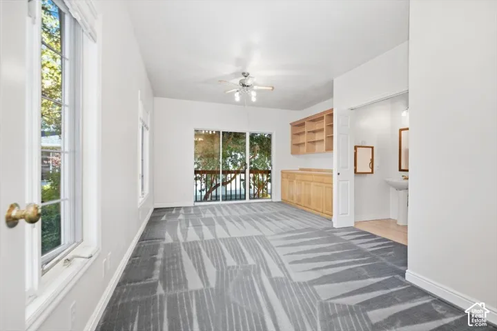 Spacious room featuring broadloom carpeting with a geometric pattern, large windows, sliding glass door access to an exterior balcony, built-in wooden cabinetry with open shelving, and an overhead ceiling fan