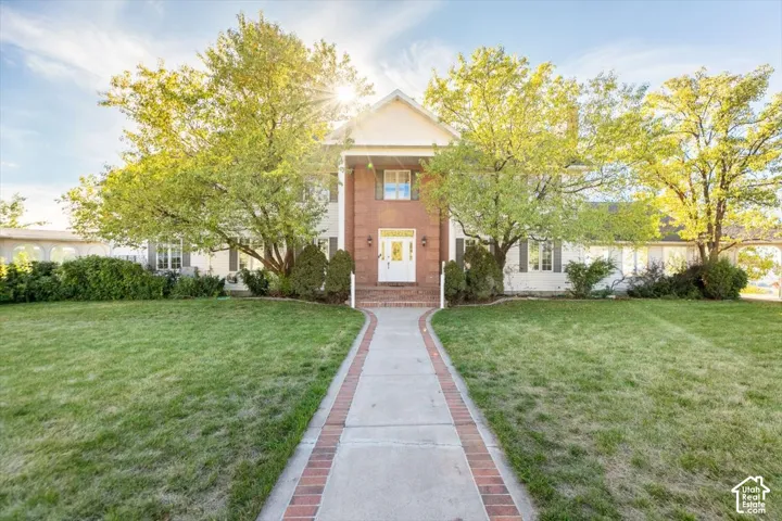 Brick facade with white columned portico, white front door, symmetrical mature trees, and a concrete paver walkway