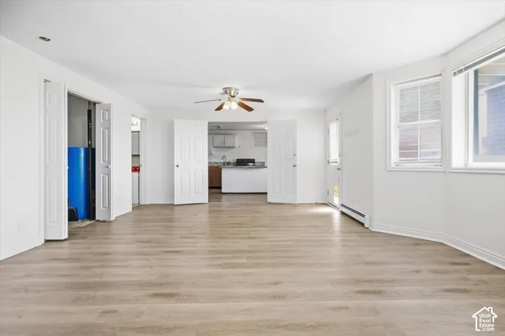 Light wood-finish flooring, white painted walls, ceiling fan with light fixture, multiple windows with blinds, and white interior doors