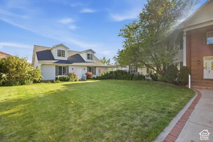 Expansive green lawn, brick-lined concrete pathway, mature trees, and a two-story residence with white siding and dormer windows