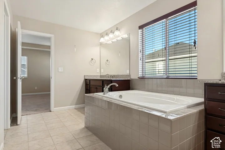 Tile flooring, built-in soaking tub with tile surround, dual vanities with granite countertops, wood-finish cabinetry, and a large window with blinds