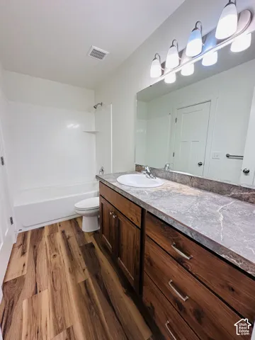 Wood-finish flooring, single vanity with a white basin sink, gray countertop, dark wood cabinetry, and a white shower-tub combination