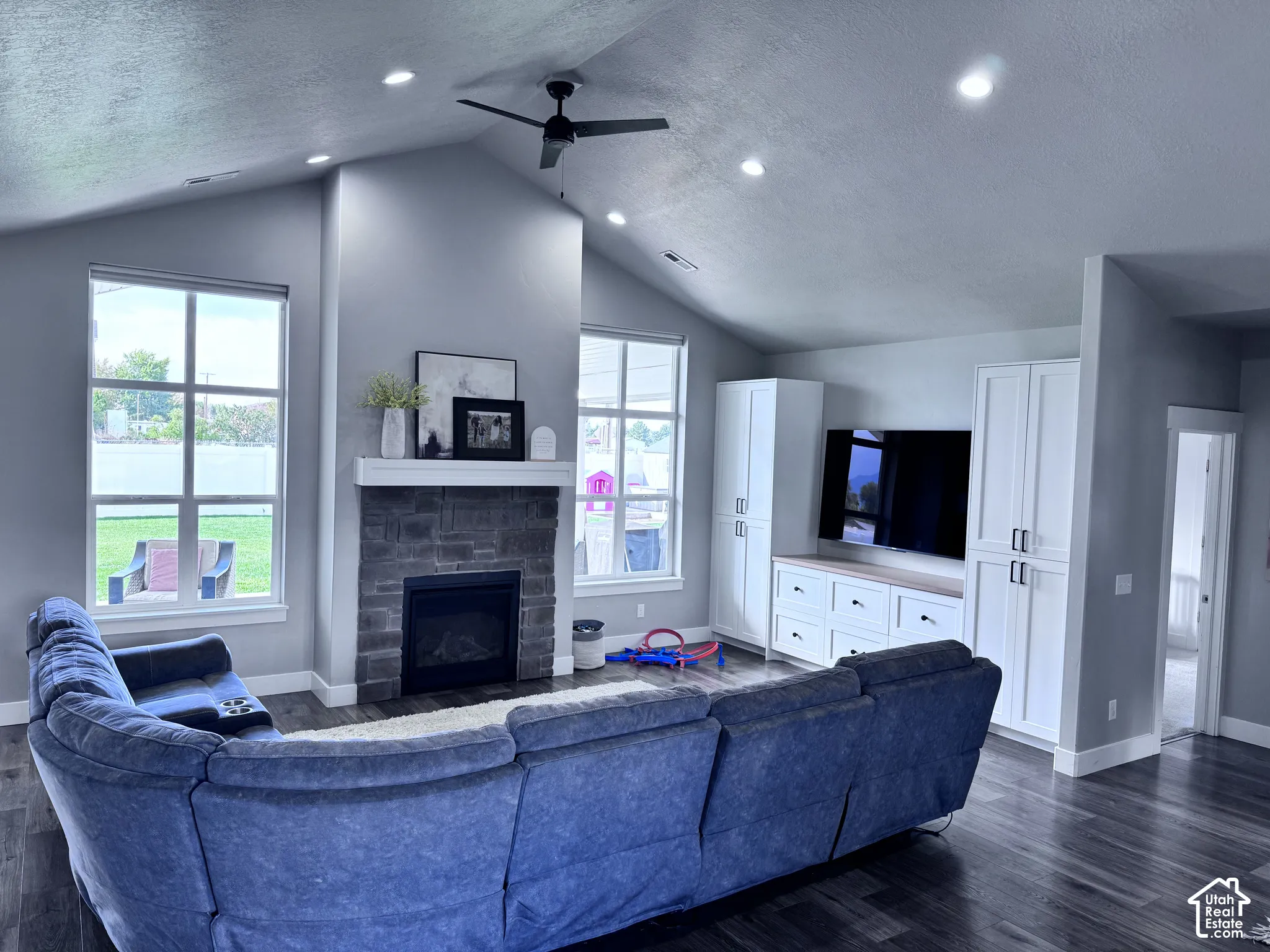 Vaulted ceiling with recessed lighting, stone-clad fireplace with mantel, built-in media cabinetry with wood-finish countertop, ceiling fan, and wood-finish flooring