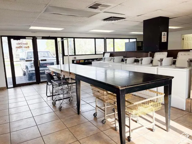 Commercial laundry facility featuring multiple washing machines, tiled flooring, and extensive natural light from storefront windows