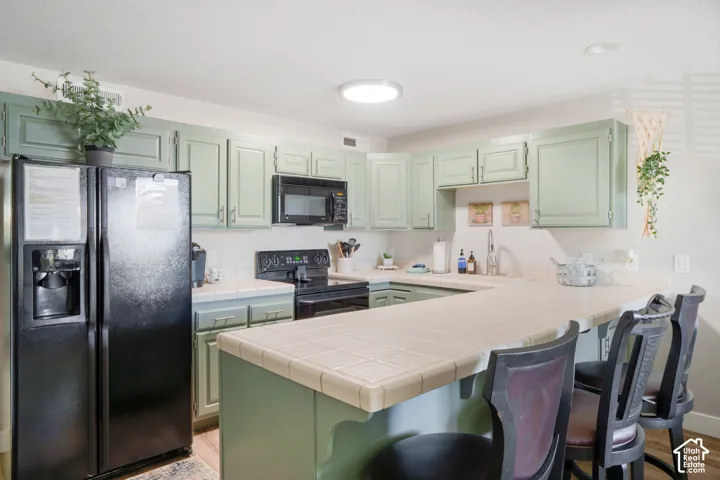 U-shaped kitchen featuring light green cabinetry, tile countertops, and a black appliance suite including a refrigerator, oven, and microwave