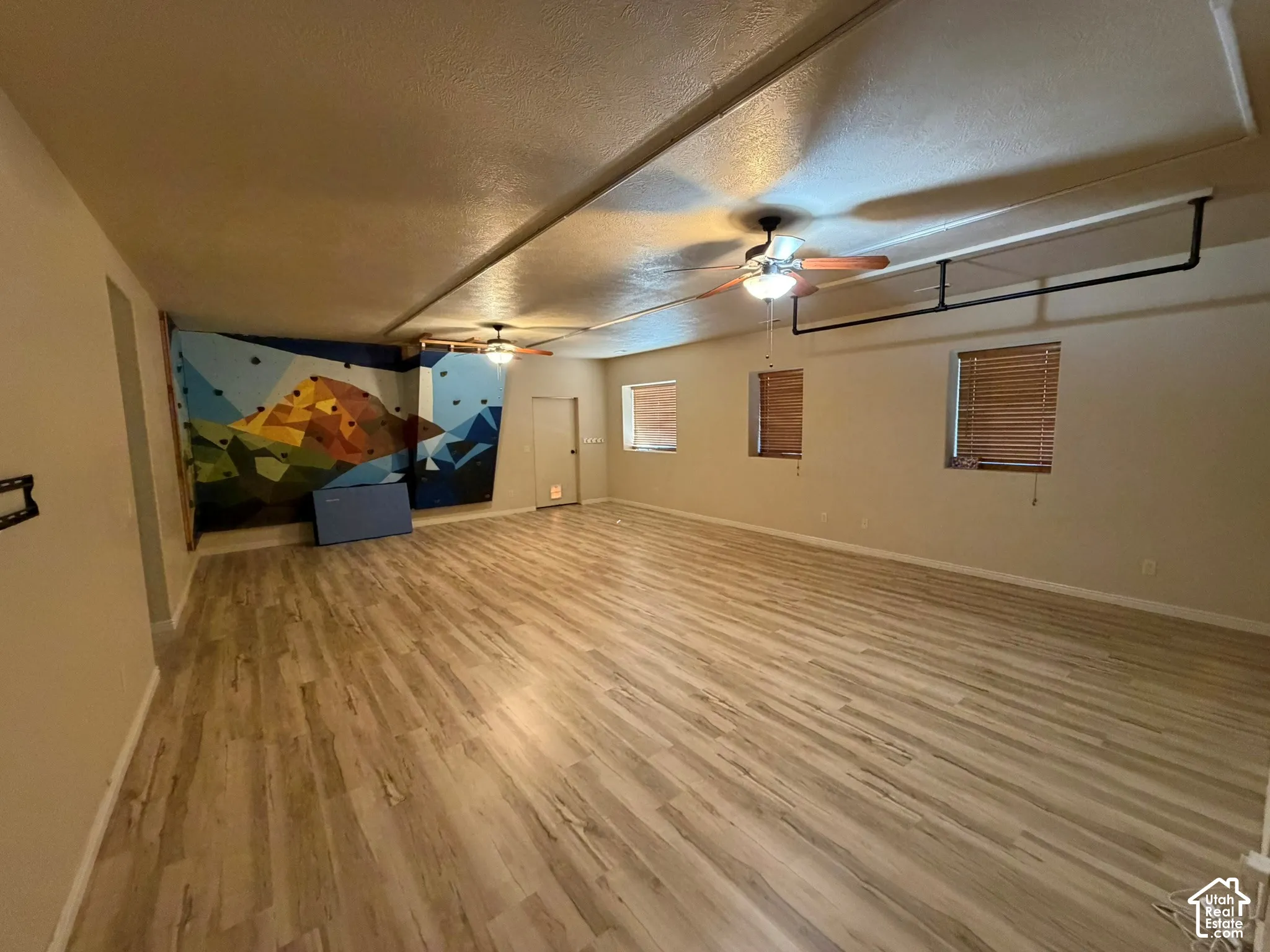 Expansive room featuring wood-finish flooring, two ceiling fans with integrated lighting, and multiple windows with blinds