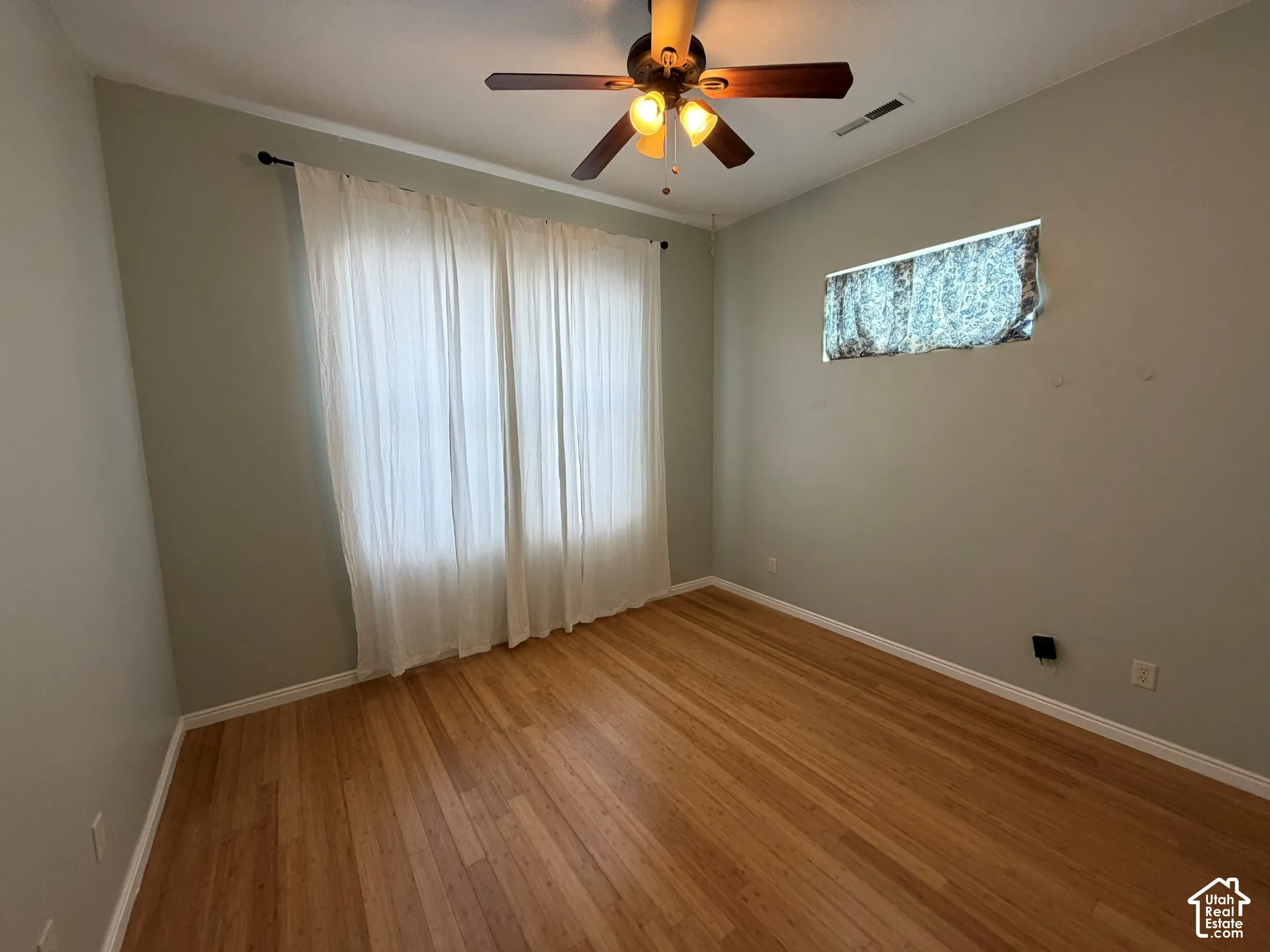 Hardwood-finish flooring, ceiling fan with integrated lighting, window with sheer curtains, and light-toned wall paint