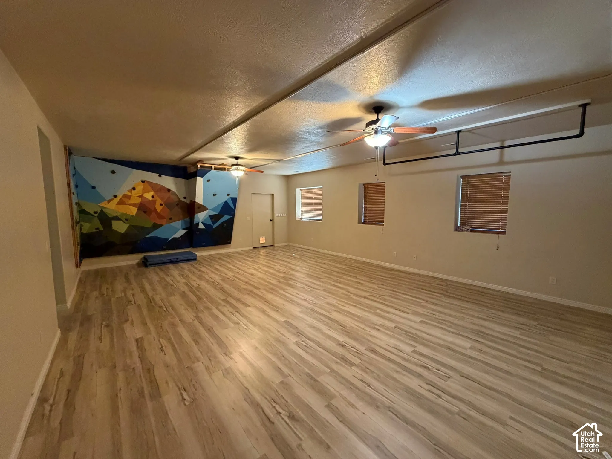 Expansive room featuring wood-finish flooring, two ceiling fans, multiple windows with blinds, and a climbing wall with handholds