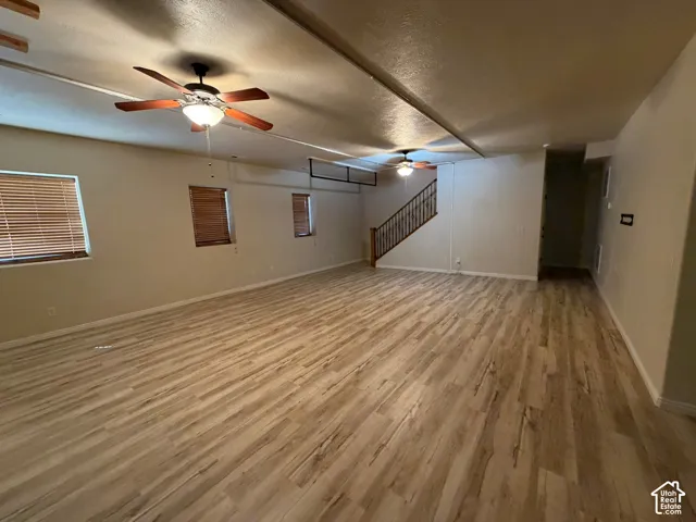 Spacious room featuring wood-finish flooring, two ceiling fans with integrated lighting, multiple windows with wood blinds, and a staircase with wood handrails and metal balusters