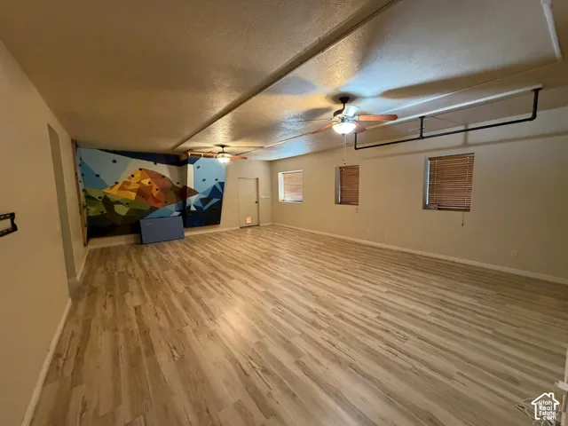 Expansive room featuring wood-finish flooring, two ceiling fans with integrated lighting, and multiple windows with blinds