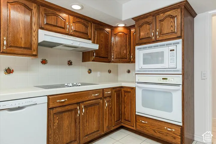 Wood-finish cabinetry with decorative hardware, white tile countertops, integrated microwave and oven, built-in electric cooktop, and white tile flooring