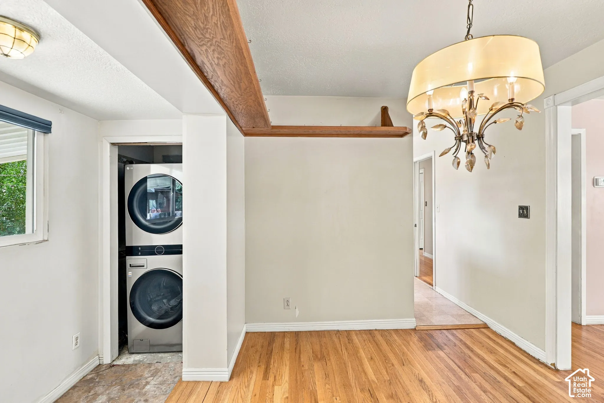 Hardwood flooring, exposed wood beam, stacked laundry appliances, chandelier light fixture, and a window with a roller shade