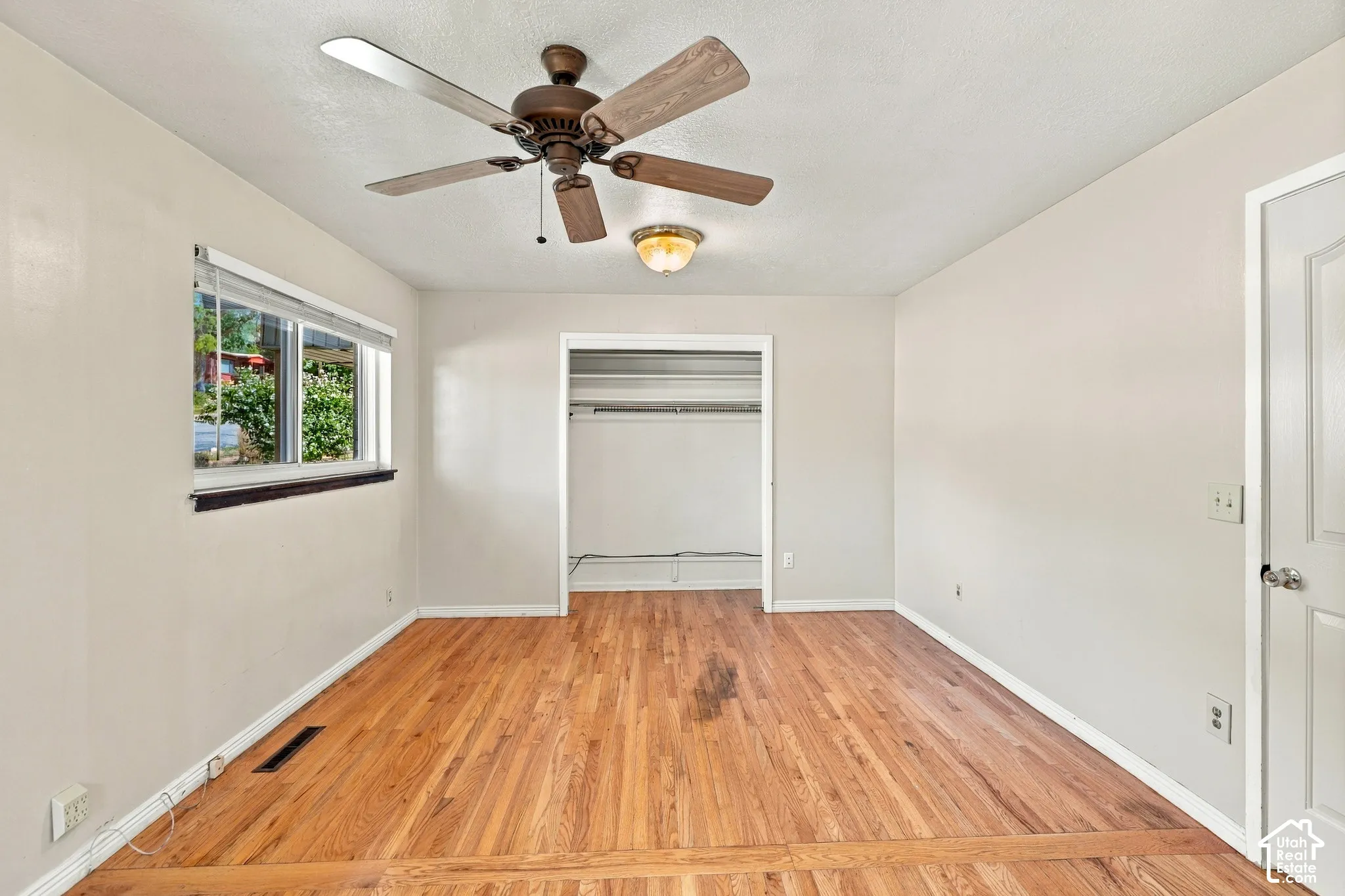 Wood-finish flooring throughout, ceiling fan with integrated lighting, recessed closet area with hanging rod, and a window with white blinds