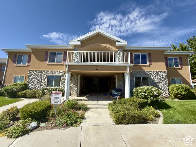 Stucco exterior with stone accents, second-story balcony with white railing, covered entryway, mature landscaping, and concrete walkways