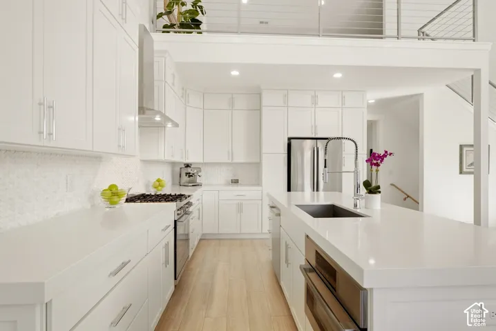 Contemporary kitchen featuring white cabinetry, integrated stainless steel appliances, a center island with sink, light wood-finish flooring, and a white tile backsplash