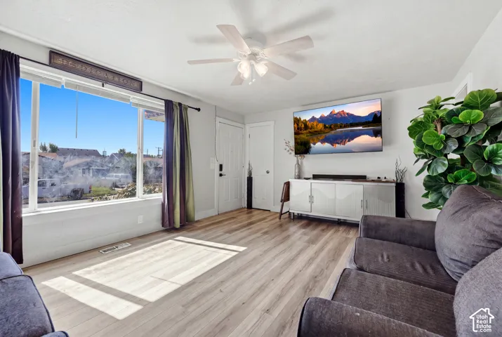 Wood-finish flooring, white ceiling fan with integrated lighting, large picture window, white interior door, and a built-in wall-mounted television
