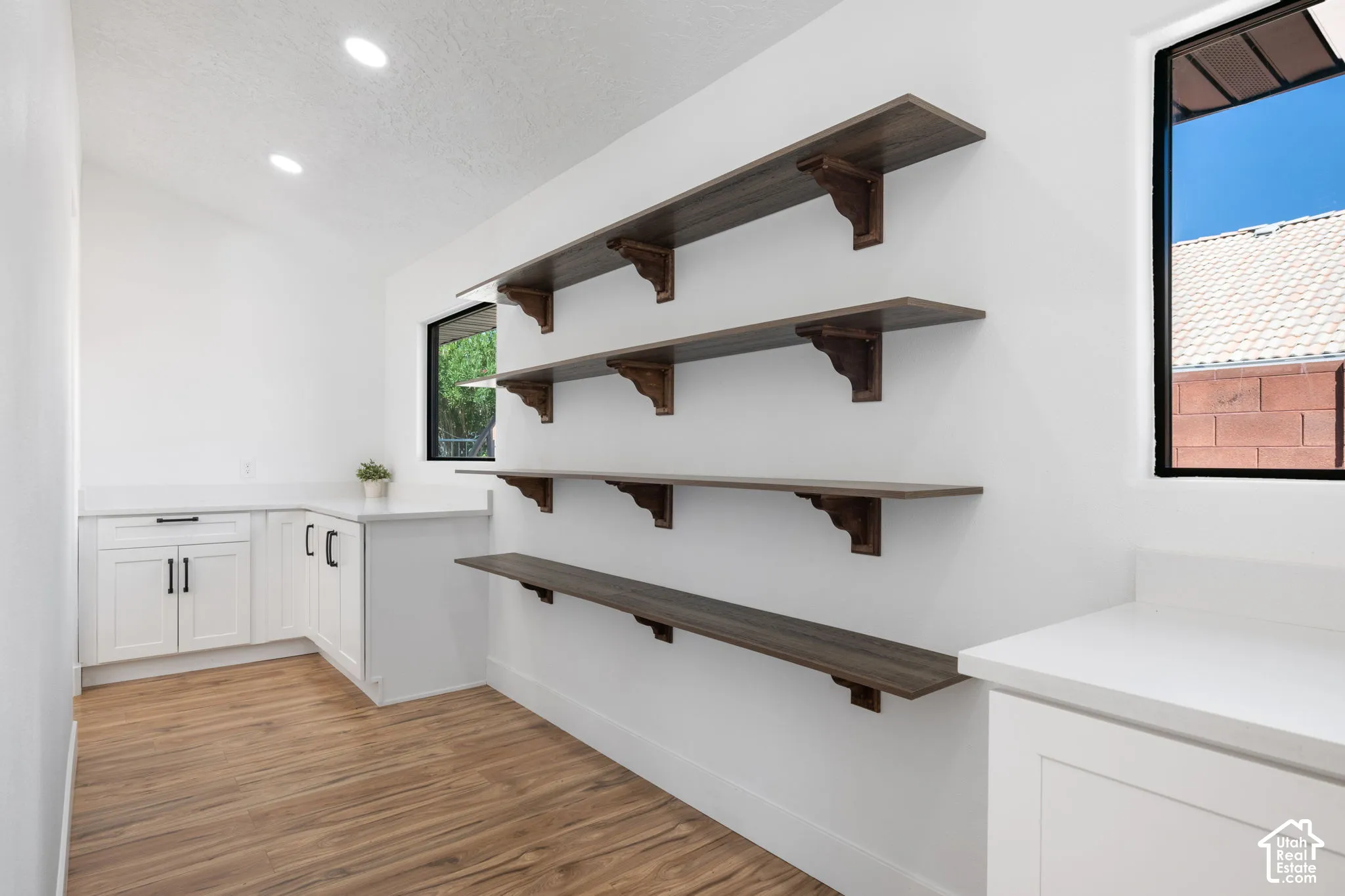 Wood-finish flooring, recessed lighting, white cabinetry with dark hardware, light-toned countertops, and multiple wood-finish floating shelves