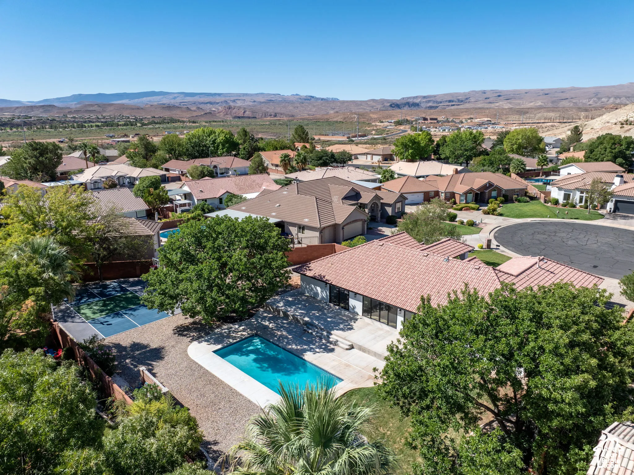 Outdoor swimming pool, fenced tennis court, mature trees, tiled roofing, and desert landscape backdrop