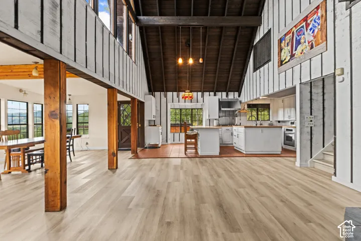 Vaulted ceiling with exposed beams, wood-finish flooring, white shiplap walls, kitchen with white cabinetry, and multiple windows
