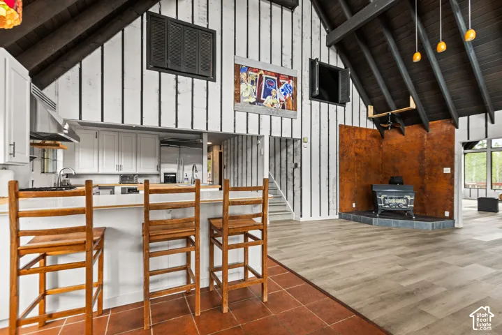 Vaulted wood-paneled ceiling, white vertical board and batten walls, wood-finish flooring, built-in kitchen cabinetry, and a hearth with a freestanding wood stove