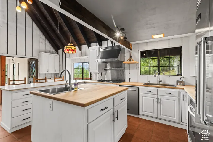 Kitchen featuring an angled ceiling with exposed wood beams, central island with an undermount sink, wood-finish countertops, white cabinetry with dark hardware, and stainless steel appliances
