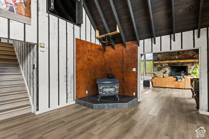 Wood-finish flooring throughout, exposed beam vaulted ceiling, wood-burning stove with tiled hearth, textured vertical wall paneling, and a stone-clad fireplace surround