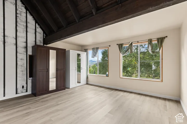 Vaulted ceiling with exposed wood beams, dual windows with wood trim, wood-finish flooring, and a textured accent wall