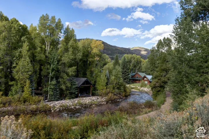 Wood-sided structures with green roofs, surrounded by dense mature trees, alongside a natural river, with distant mountains