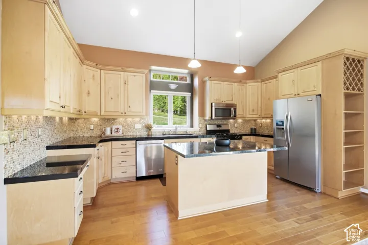 Kitchen with light wood-finish cabinetry, dark countertops, a mosaic tile backsplash, stainless steel appliances, and a central island