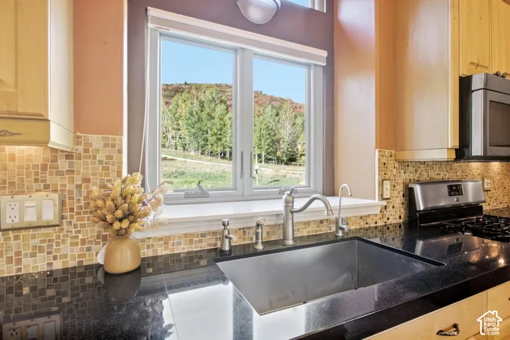 Stainless steel sink with a gooseneck faucet, black countertop, mosaic tile backsplash, light wood cabinetry, and a window with natural scenery