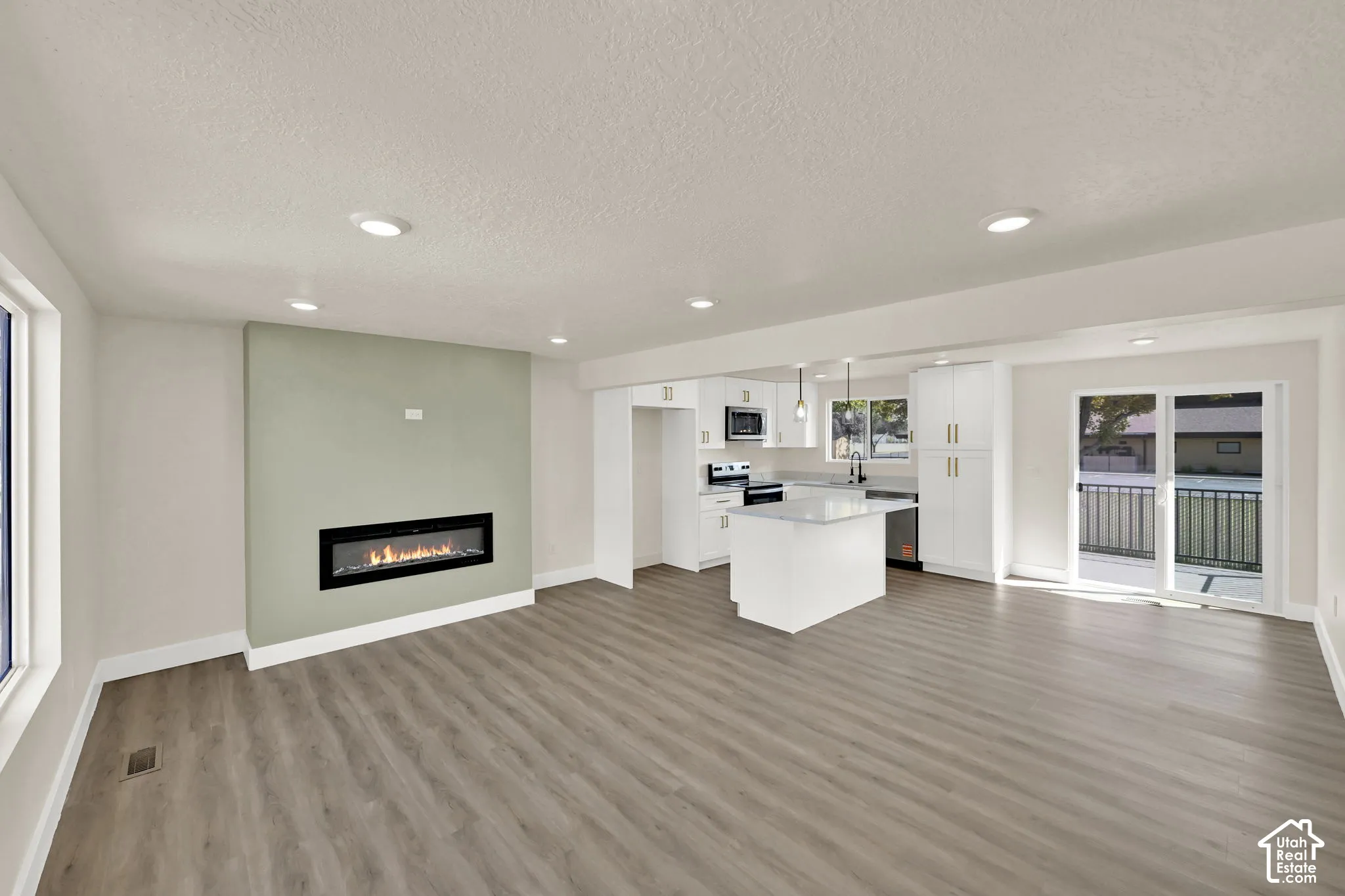 Open-concept living area featuring wood-finish flooring, a linear electric fireplace, recessed lighting, a kitchen island with pendant lighting, and sliding glass doors
