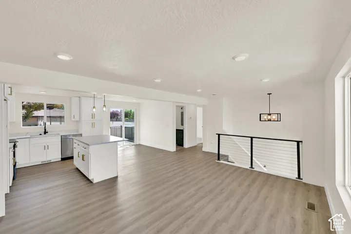 Open-concept living area with wood-finish flooring, a contemporary kitchen featuring white cabinetry and a central island, stainless steel appliances, and a modern stair railing with cable infill