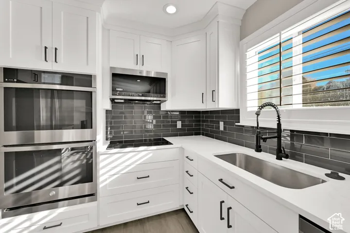 White shaker-style cabinetry with dark hardware, stainless steel double wall ovens, glass subway tile backsplash, white countertops, and a stainless steel undermount sink with a black pull-down faucet
