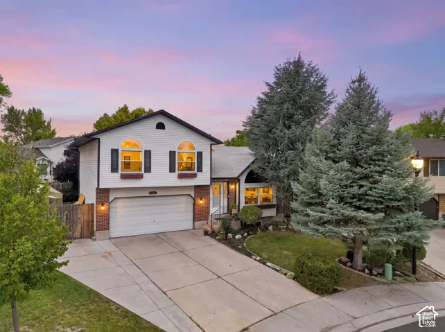 Split-level residence featuring a two-car garage, arched windows with shutters, brick accents, and mature evergreen landscaping