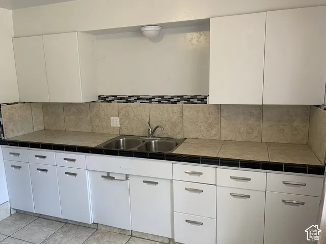 White upper and lower cabinetry, dual basin stainless steel sink, tile countertops, and tile backsplash with decorative accent