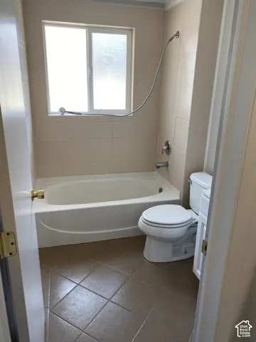 White tub and shower combination with tiled surround, frosted window, and tile flooring