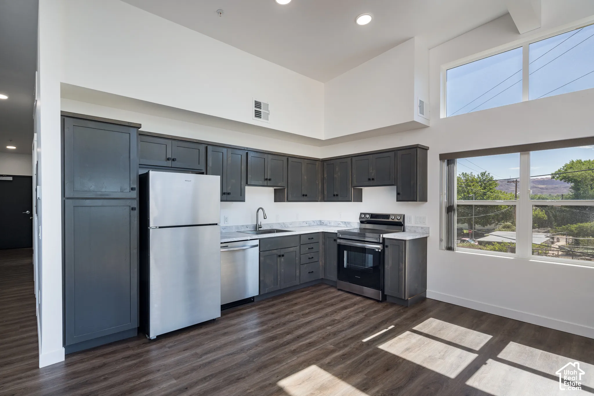 Kitchen featuring dark wood-finish cabinetry, stainless steel appliances, light-colored countertops, wood-finish flooring, and high ceilings with upper-level windows