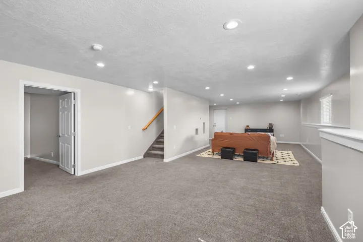 Expansive carpeted area with recessed lighting, a window with blinds, and a staircase featuring a wood handrail