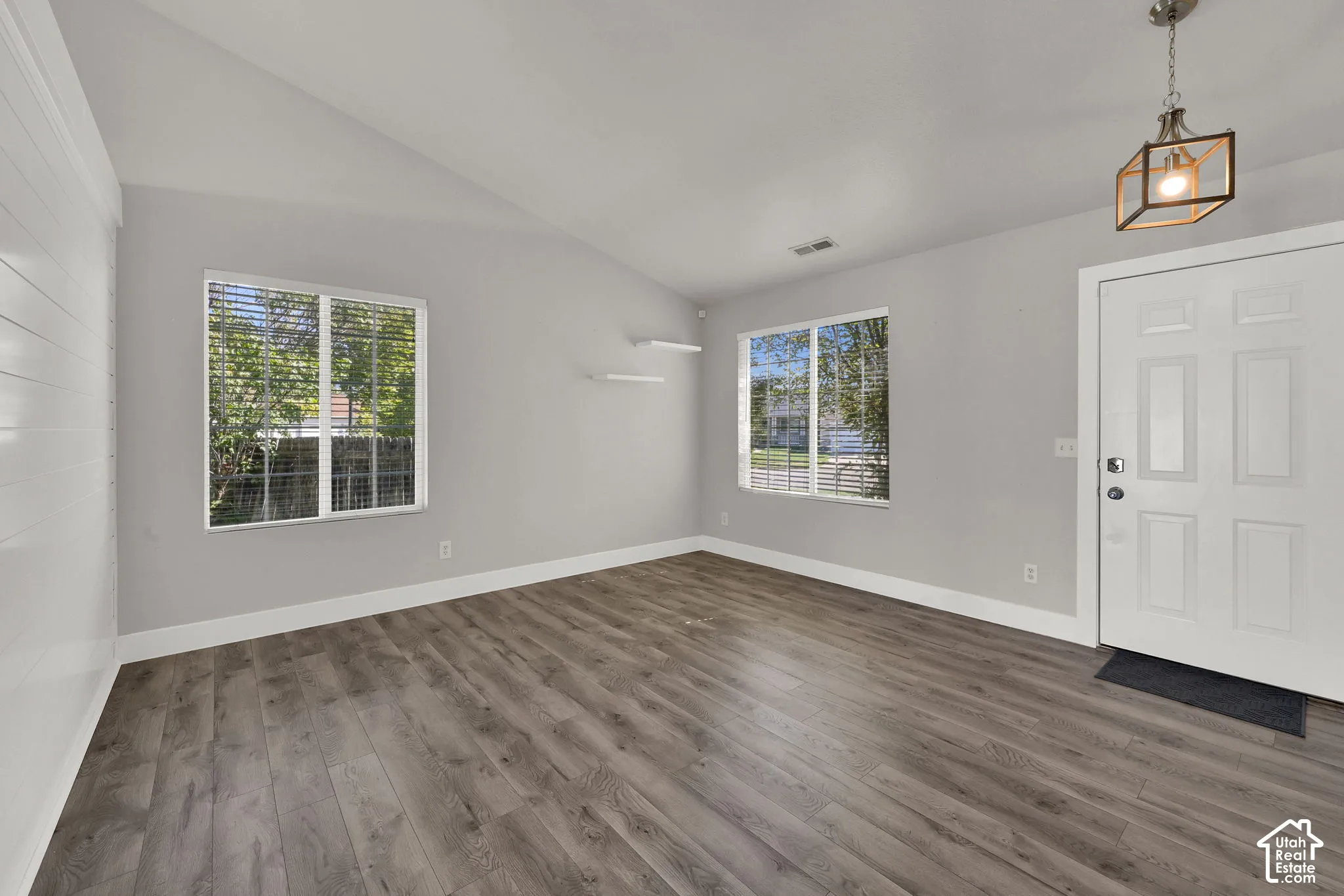 Vaulted ceiling, wood-finish flooring, two windows with blinds, panel door, and a geometric pendant light fixture