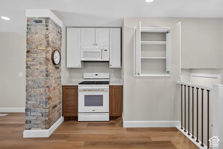 Kitchen featuring a gas range, microwave, light-toned upper cabinetry, wood-finish flooring, and an exposed brick wall