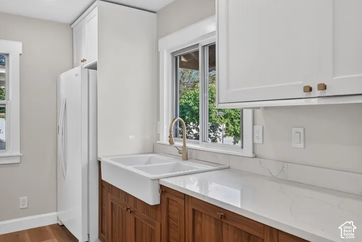 White shaker-style upper cabinetry, wood-finish lower cabinetry, dual basin farmhouse sink, brushed brass faucet, and white countertop